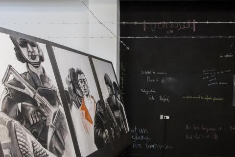 Trois dessins avec différentes personnes dessus. Au fond de la photo, un mur noir avec des écritures manuscrites. 